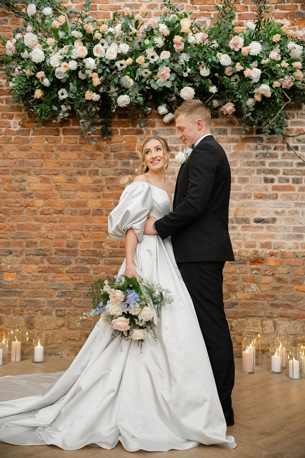 bride and groom under floral installation in front of brick wall