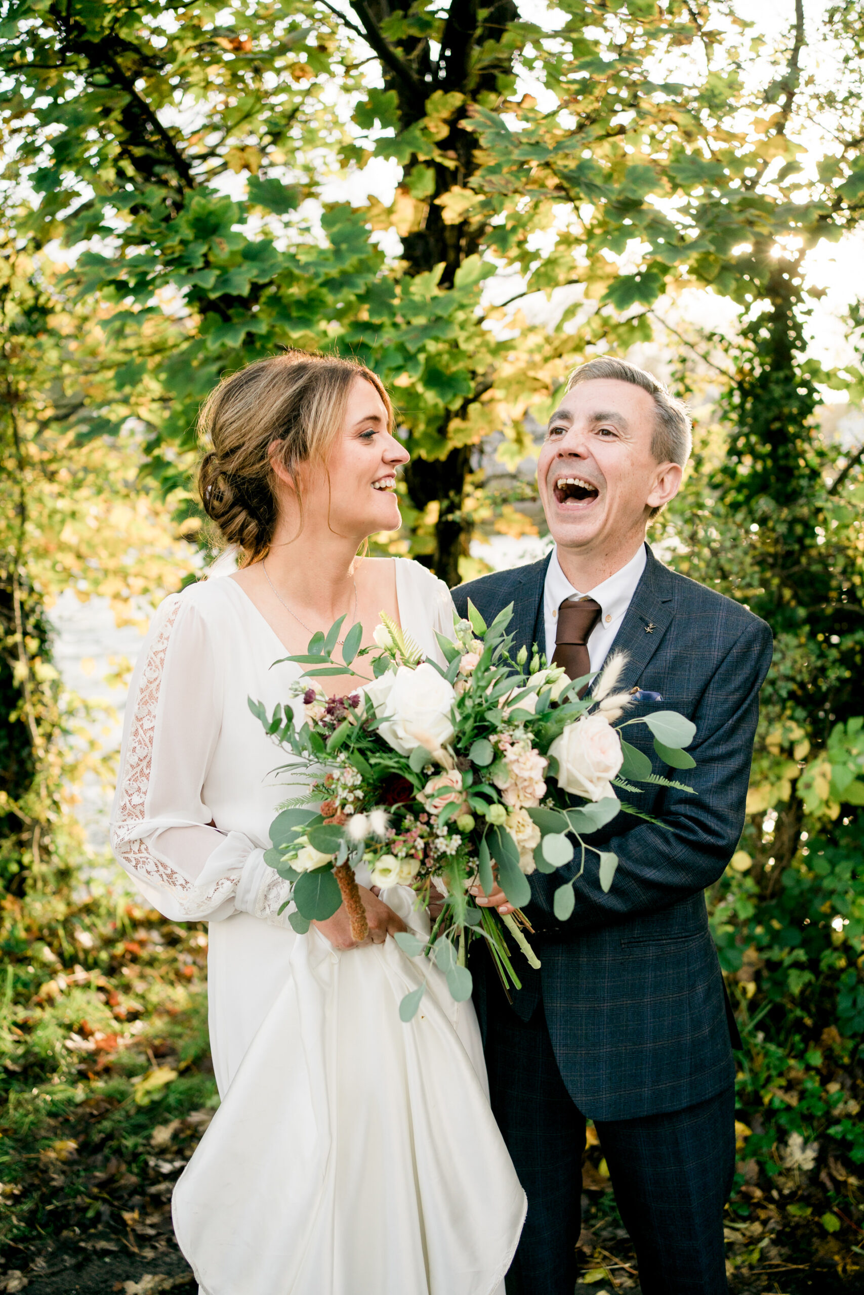 bride and groom laughing in front of tree lined river