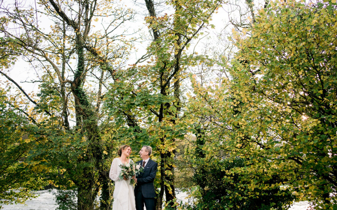 bride and groom in front of trees and river at the swan hotel newby bridge