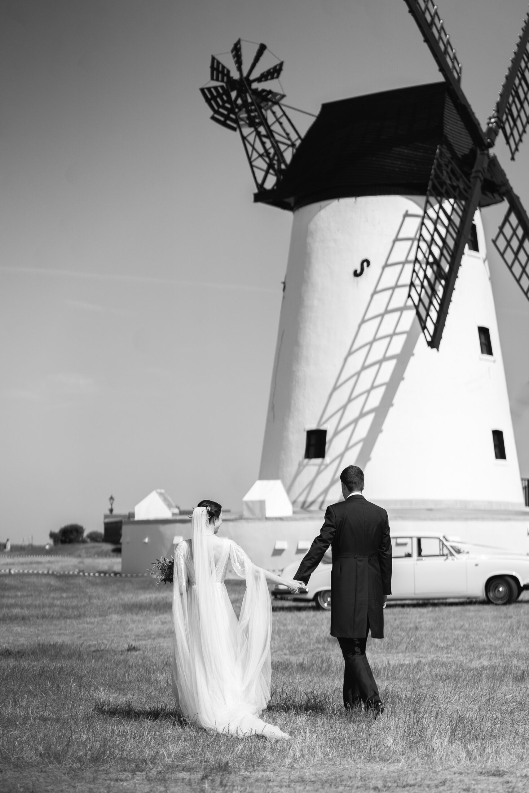 bride and groom photographed from behind walking towards their wedding car, with Lytham Windmill in the background