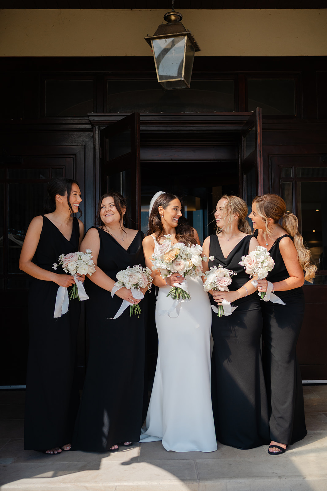 bride with cream and blush bouquet giggles with bridesmaids in black dresses, on the steps at the Clifton arms hotel in lytham