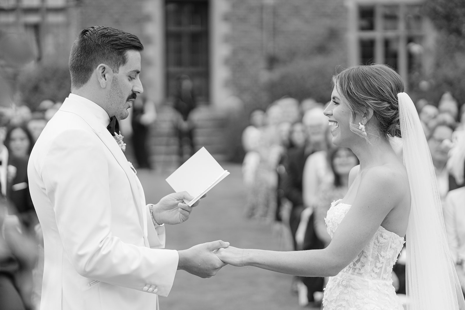 groom holds brides hand as he reads his vows for the card, she smiles widely