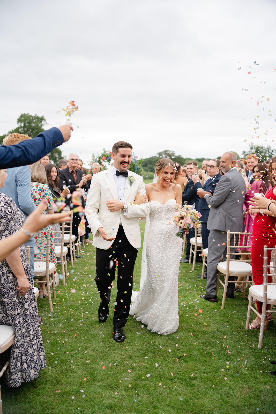 bride and groom walk down the grassy aisle with guests throwing confetti and cheering