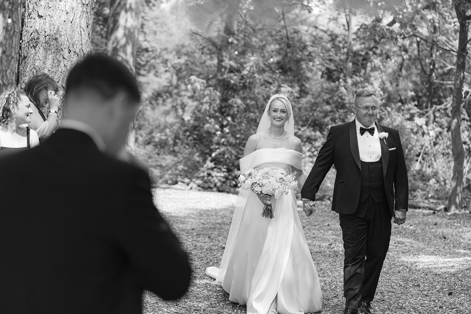 groom wipes away tears as bride and her dad walk down the aisle